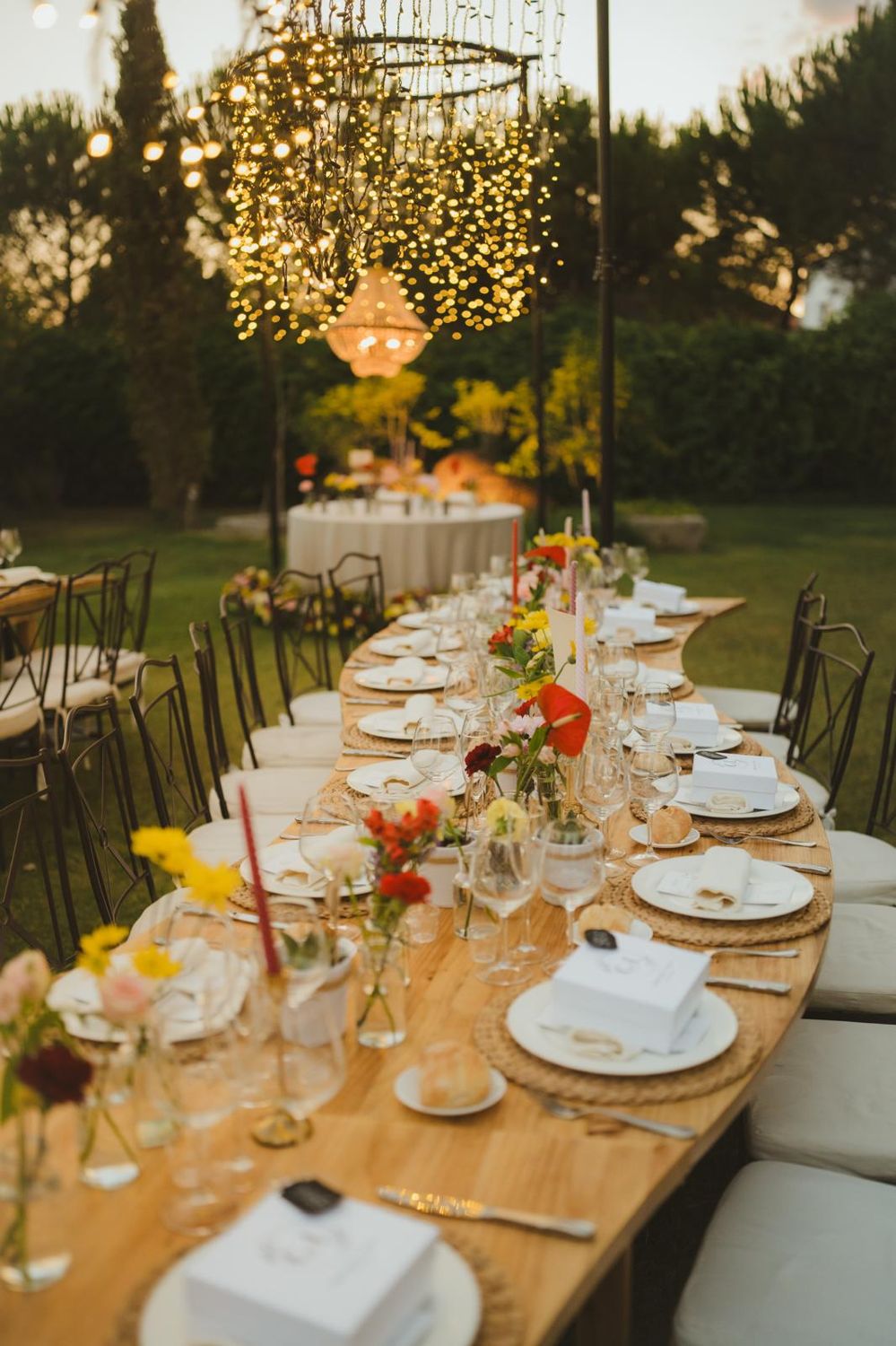 Mesa larga con velas de colores, flores y lámpara de cristal al fondo