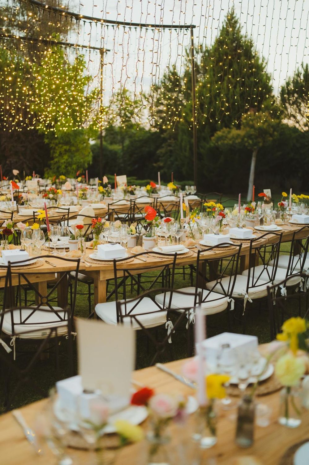 Largas mesas de madera con flores bajo cortinas de luces en el jardín